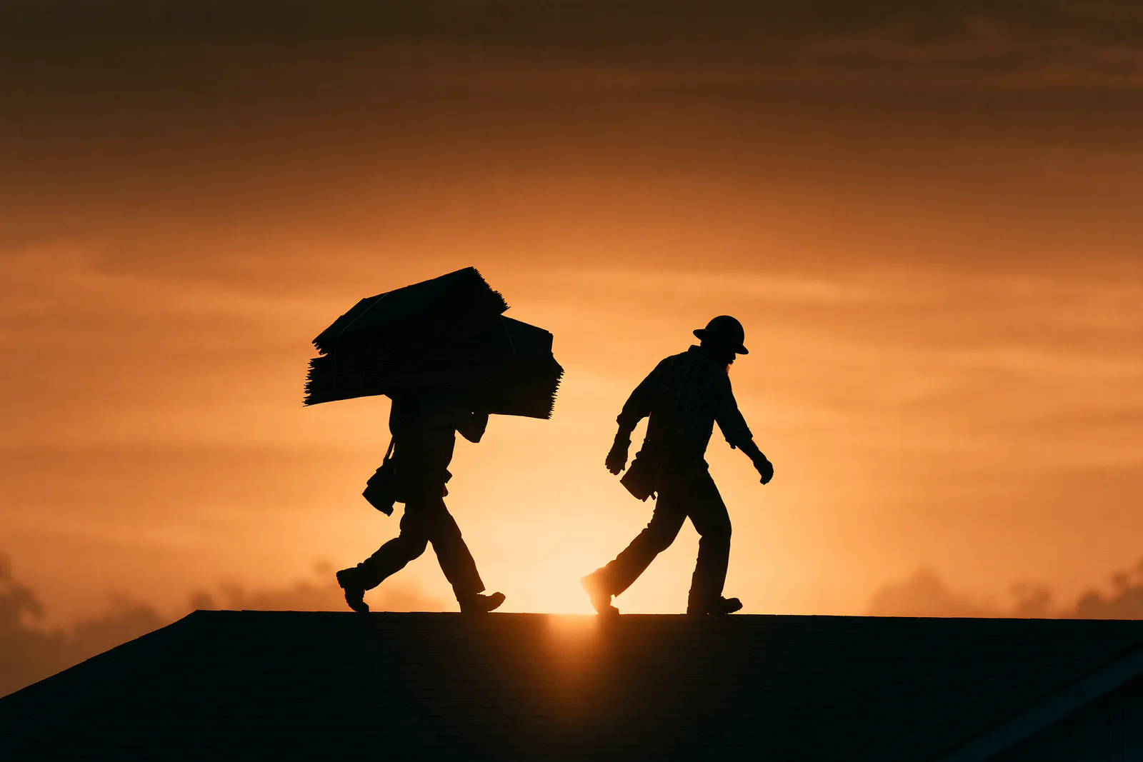 Two roofers silhouetted against a sunset walk along a roof ridge one carrying shingle bundles